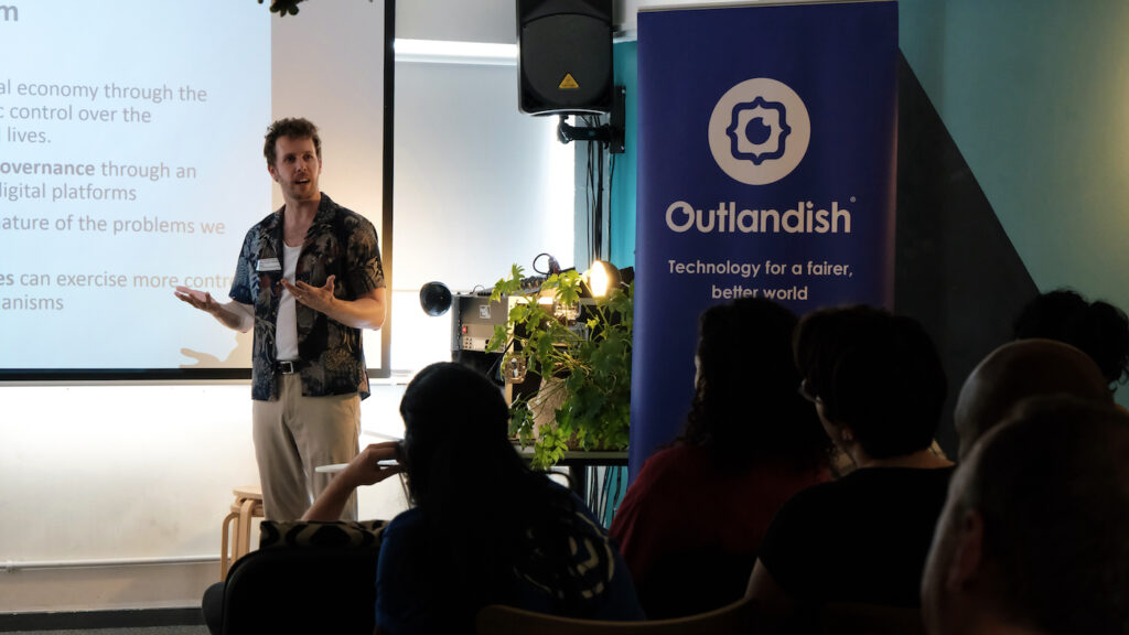 Man presenting and standing next to a roll-up banner that reads "Outlandish. Technology for a fairer, better world."