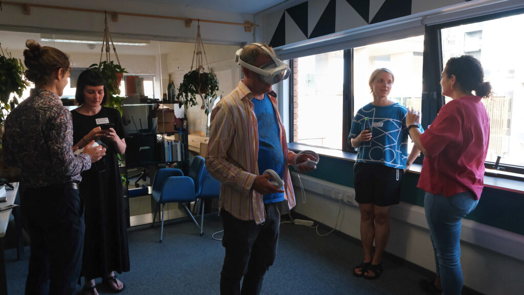 Five people in a room. Two pairs talking to each other at opposite end. One person in the middle wearing a VR headset.