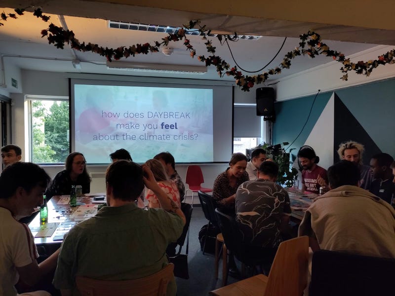 A zoomed out image of SPACE4 with two tables of people playing daybreak. A presentation in the back of the room reads "How does Daybreak make you feel about the climate crisis"