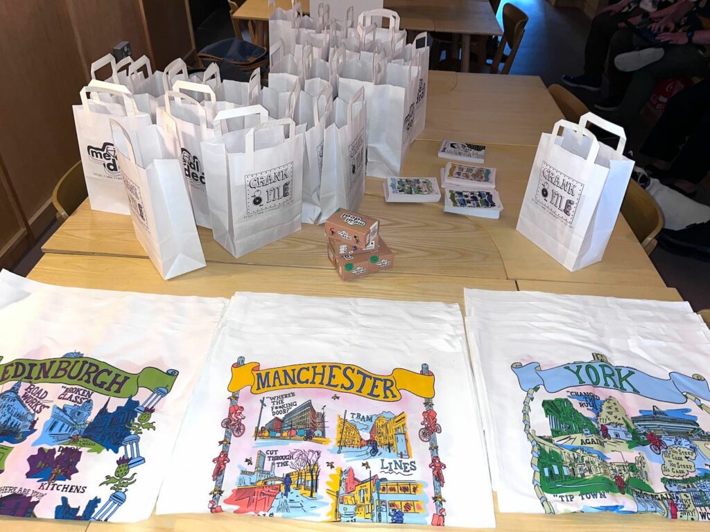 Picture of merchandise: lots of white bags with 'crank file' written on them. And illustration tea towels of Edinburgh, Manchester and York.