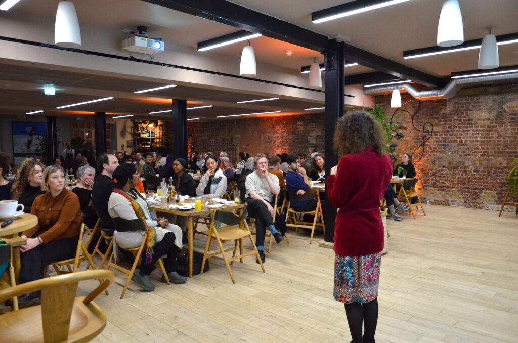 Photograph of Miri from Disrupt presenting to a seated audience at dining tables